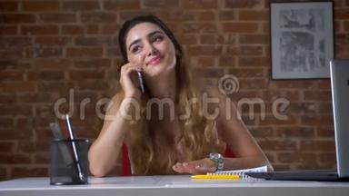 可爱的白种人女孩在办公室里坐在靠近笔记本电脑的桌面上轻松地用手机说话，工作轻松